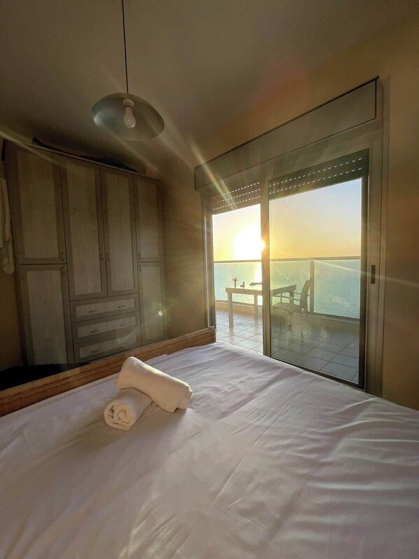 Quarto de hotel com cama, guarda-roupa, uma grande janela com vista para o mar e uma mesa exterior.