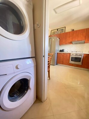 A laundry area with stacked washing machines and a kitchen with wooden cabinets and stainless steel appliances. - Krystal View Family 2 parking Bomb Shelter (Bat Yam)