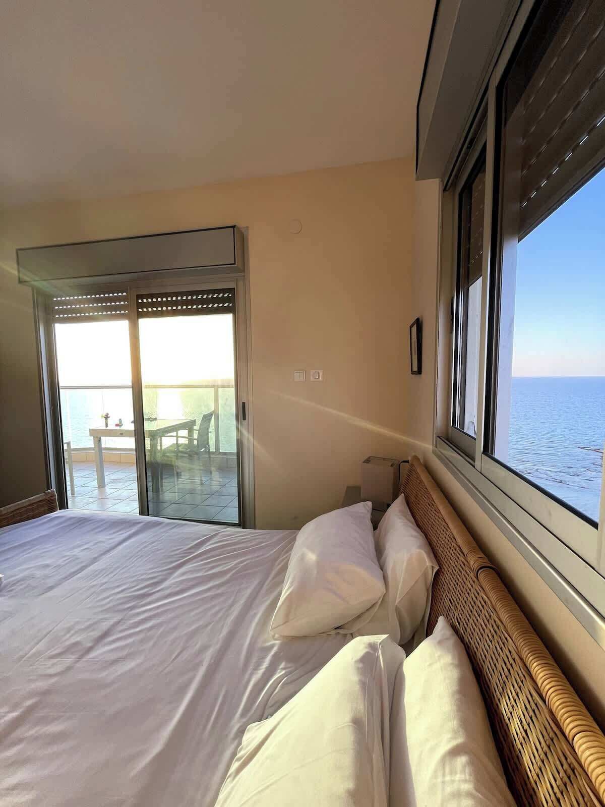 A hotel room with a bed, a window with a view of the sea, and a sliding glass door leading to an outdoor area.