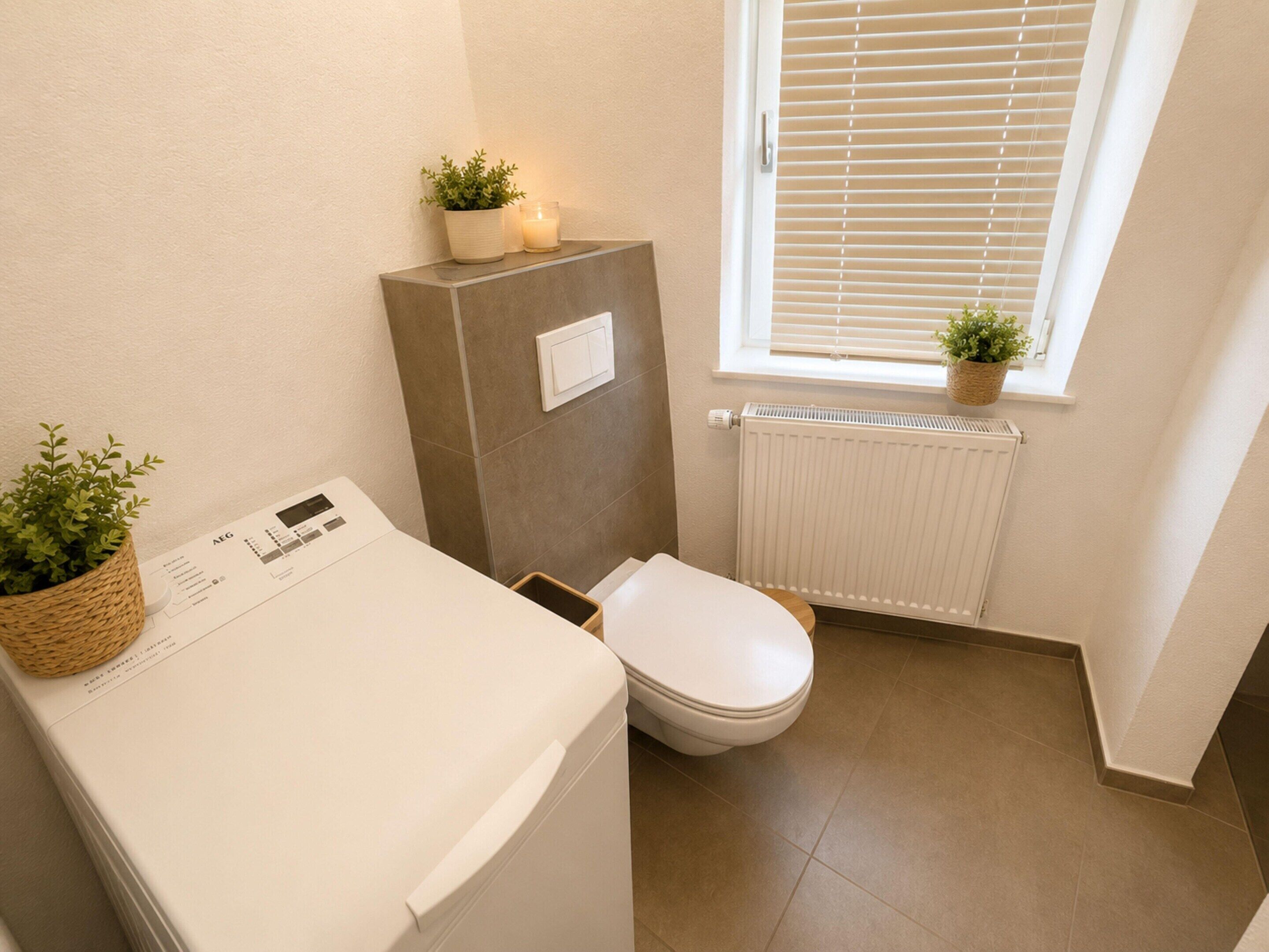 Et lille badeværelse med vaskemaskine, toilet, radiator og et vindue med persienner.