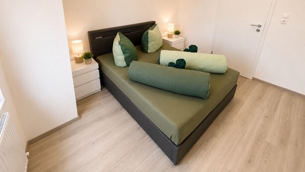 Cama bem arrumada com roupa de cama verde, uma luminária de cabeceira e uma pequena planta sobre um criado-mudo branco.