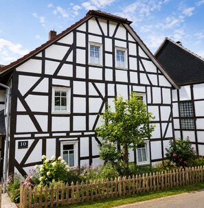 Ett traditionellt hus med korsvirkesfasad, ett nummer ’10’ på dörren, ett staket av träspjälor och ett träd på framsidan av tomten.