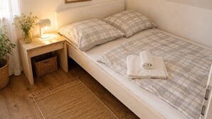 Ein Zimmer mit einem Einzelbett, geneigter Decke, einem Nachttisch mit Lampe, einem gerahmten Pflanzenbild und einem gewebten Teppich.