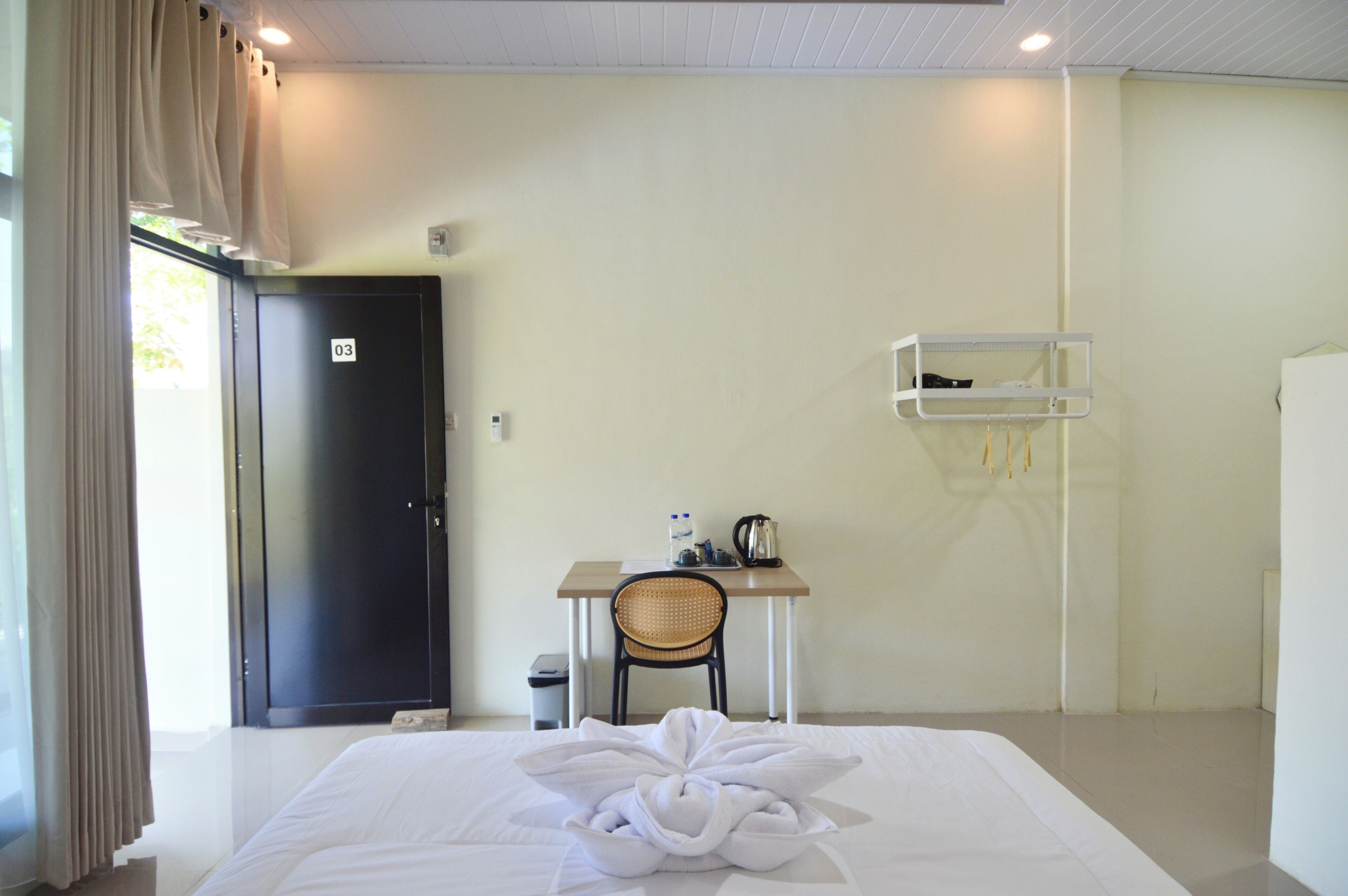 A hotel room with a bed, a desk with a chair, a small shelf, and a door marked with the number 03.