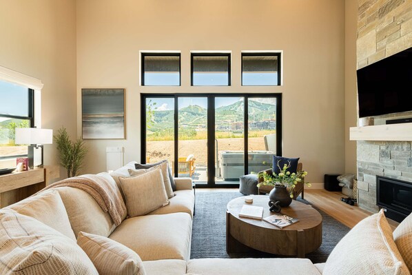 Una sala de estar moderna con sof á, mesa de centro y chimenea. Se observan grandes ventanales con vistas a un paisaje montañoso.