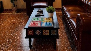 A room with two leather sofas, a coffee table with a board game, a window with plants, and a lamp. - Coastal Palm (Alibag)