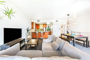 Ein modernes Wohnzimmer mit Flachbildfernseher, einer Couch, einem Esstisch und einer Küche im Hintergrund.