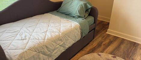 Ein Einzelbett mit gesteppter Bettdecke und Kissen, ein runder Teppich mit Blumenmuster und ein Fenster mit Jalousien.