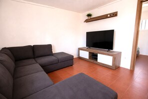 Ein modernes Wohnzimmer mit einem Ecksofa, einem Flachbildfernseher auf einem Holzständer und einer kleinen Pflanze auf einem Regal.