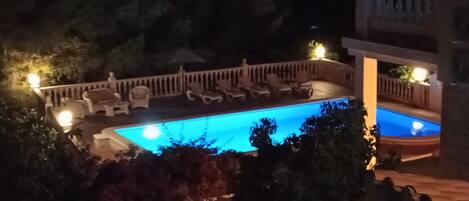Vista nocturna de una piscina con sillones de descanso y una zona de estar cubierta.