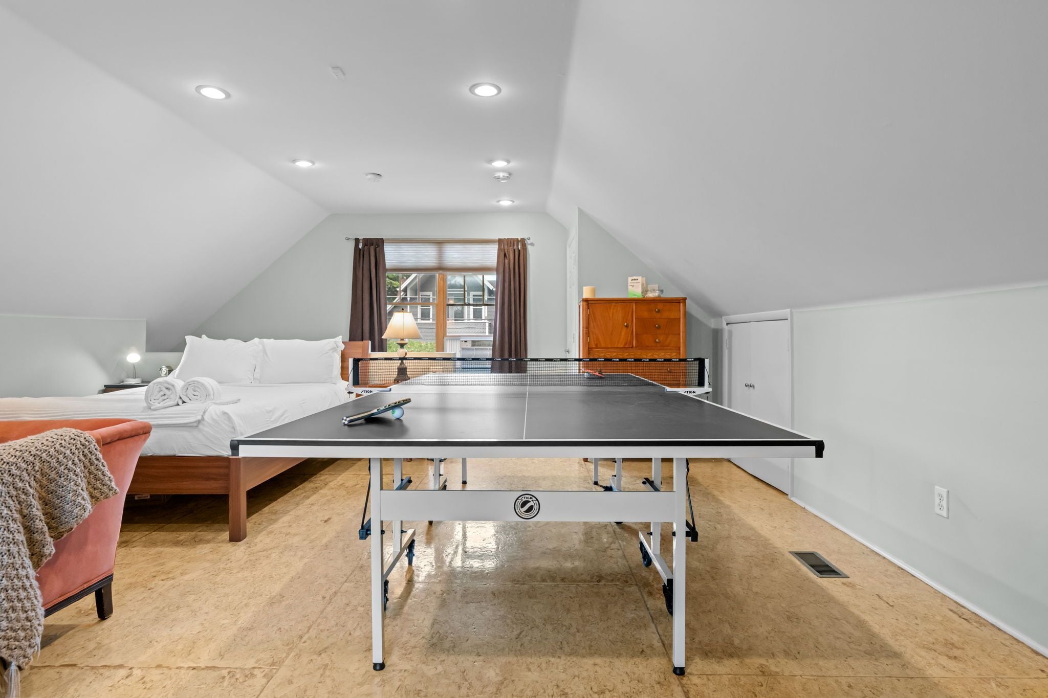A room with a ping pong table, a bed, a desk, and a window with curtains.