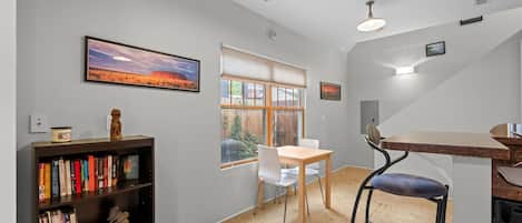 A modern kitchen with a dining area, a bookshelf, and a window with a view of a building.