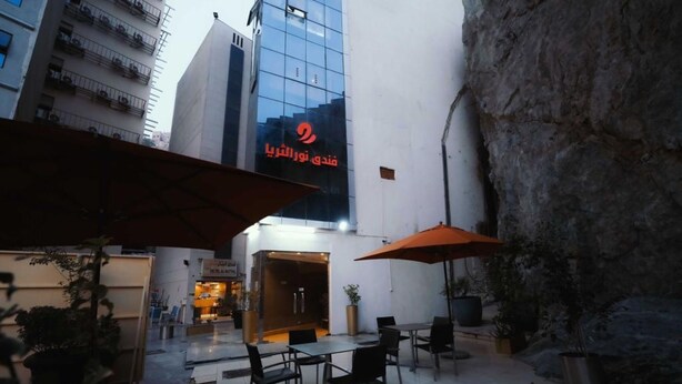An outdoor seating area with tables and chairs, a large rock wall, and a building with a glass facade in the background. - SElaf Almaqam Nour (Makkah)
