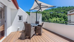 Ein Balkon mit Tisch und Stühlen, ein weißes Sonnensegel und Blick auf einen grünen Hügel.