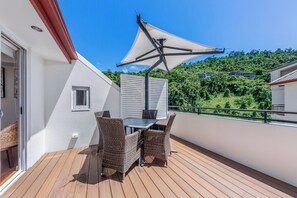 Ein Balkon mit Tisch und Stühlen, ein weißes Sonnensegel und Blick auf einen grünen Hügel.