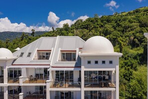 A modern multi-story building with large windows and a flat roof, surrounded by lush greenery and hills. - Whitsunday Blue Penthouse (Airlie Beach)