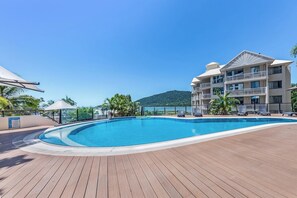 En swimmingpool med træterrasse, en bygning med flere etager og altaner, samt udsigt til et bjerg.