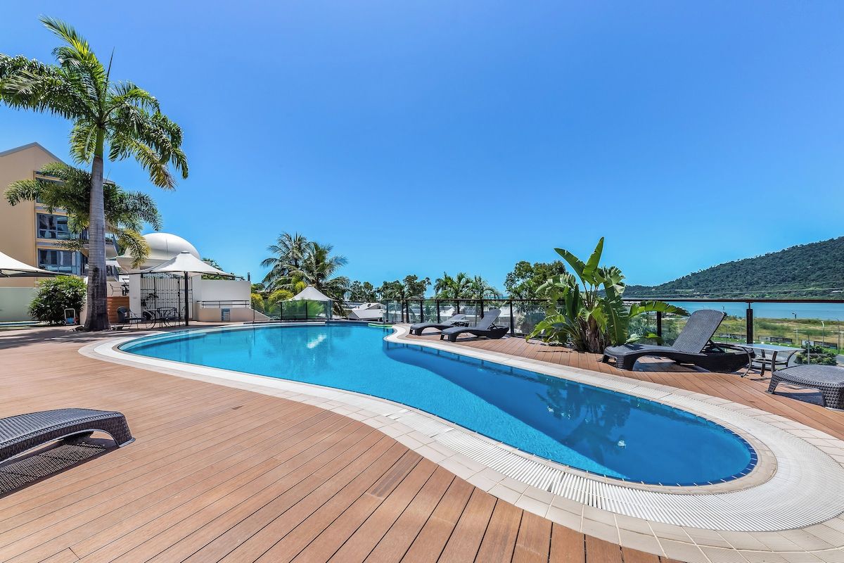 Une piscine entourée de chaises longues et de palmiers, elle-même bordée d’une terrasse en bois et d’un bâtiment surmonté d’un dôme.