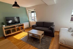 A modern living room with a flat-screen TV, a dark sofa, a wooden coffee table, and a window with a view. - Cath' Home - Studio on Place de la Cathédrale (Colmar)
