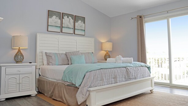 A bedroom with a bed, nightstands, lamps, and a view of the outdoors. - 🌊Coastal Townhouse | Pool & Tennis Court | Walk2Beach | Barrier Dunes 542 - 55 (Cape San Blas)