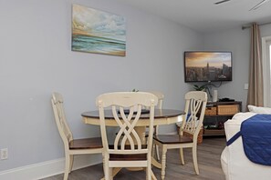 Habitación de hotel con comedor que incluye una mesa redonda y cuatro sillas, una cama con colcha azul, un televisor en la pared y un cuadro con escena de playa.