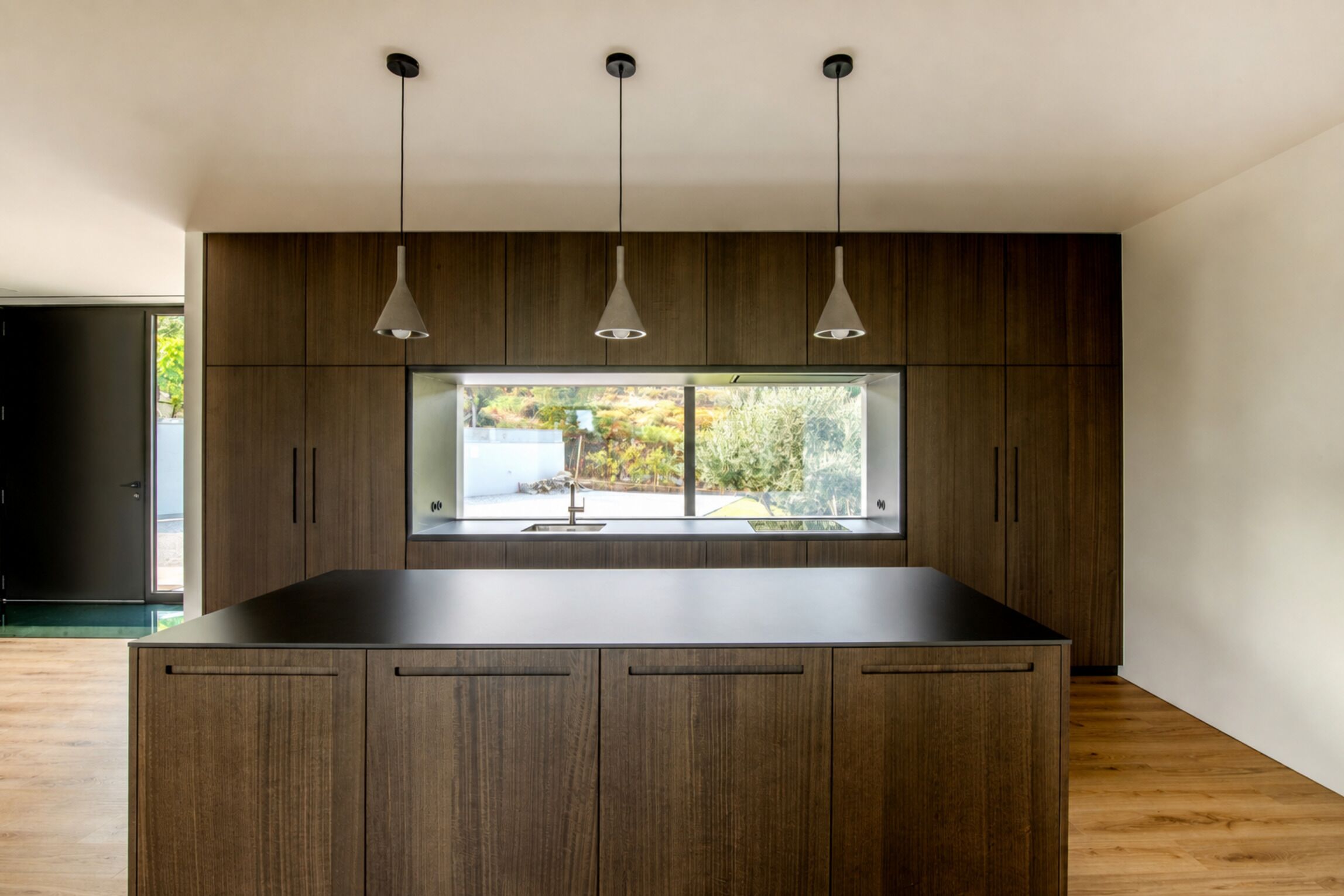 A modern kitchen with a central island, pendant lights, and a large window overlooking a garden.