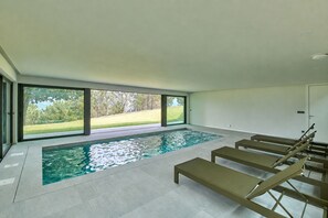 Ein moderner, überdachter Schwimmbereich mit großen Glaswänden, gefliestem Boden und einigen Liegestühlen.