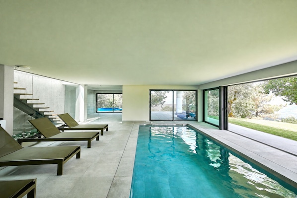 Ein moderner, überdachter Pool mit Glaswänden, Liegestühlen und Blick ins Freie.