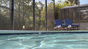 En swimmingpool med to liggestole og et overdækket område, omgivet af træer.