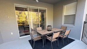 Um alpendre coberto com mesa e cadeiras, uma porta de correr e uma janela com persianas.