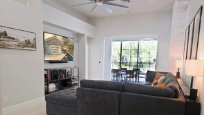 Uma sala de estar moderna com um sofá seccional cinza, uma TV de tela plana na parede e uma área de jantar com mesa e cadeiras de vidro.