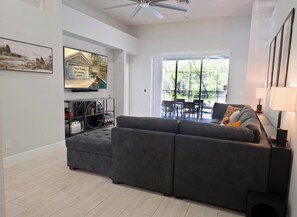 A modern living room with a grey sectional sofa, a flat-screen TV mounted on the wall, and a dining area with a glass table and chairs. - Peaceful & Modern Retreat - Pool & Themed Rooms! (Davenport)