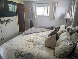 Ein Schlafzimmer mit einem Bett, Kissen, einem Nachttisch, einer Lampe, einem Fenster mit Vorhängen und einer Wandablage mit Gegenständen.