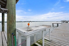 Eine Pier mit Waschbecken und Gartenschlauch, einer Holzterrasse und Blick auf Wasser sowie entfernte Gebäude.