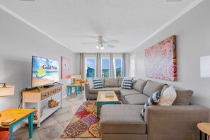 Ein Wohnzimmer mit einer Couch, einem Fernseher, einem Deckenventilator und einem großen Fenster mit Vorhängen.