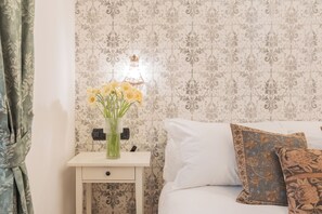 Un dormitorio con cama, una mesita de noche con un florero y papel tapiz estampado.