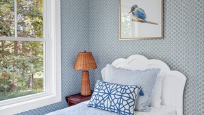 Une chambre à coucher avec un lit, un tableau encadré représentant un oiseau, une lampe et une fenêtre donnant sur des arbres.