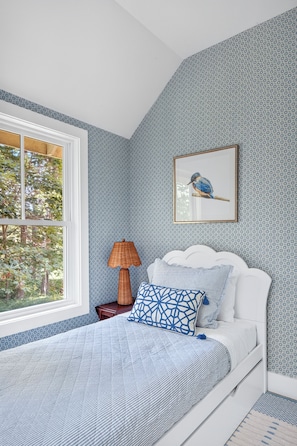 A bedroom with a bed, a framed picture of a bird, a lamp, and a window with a view of trees.