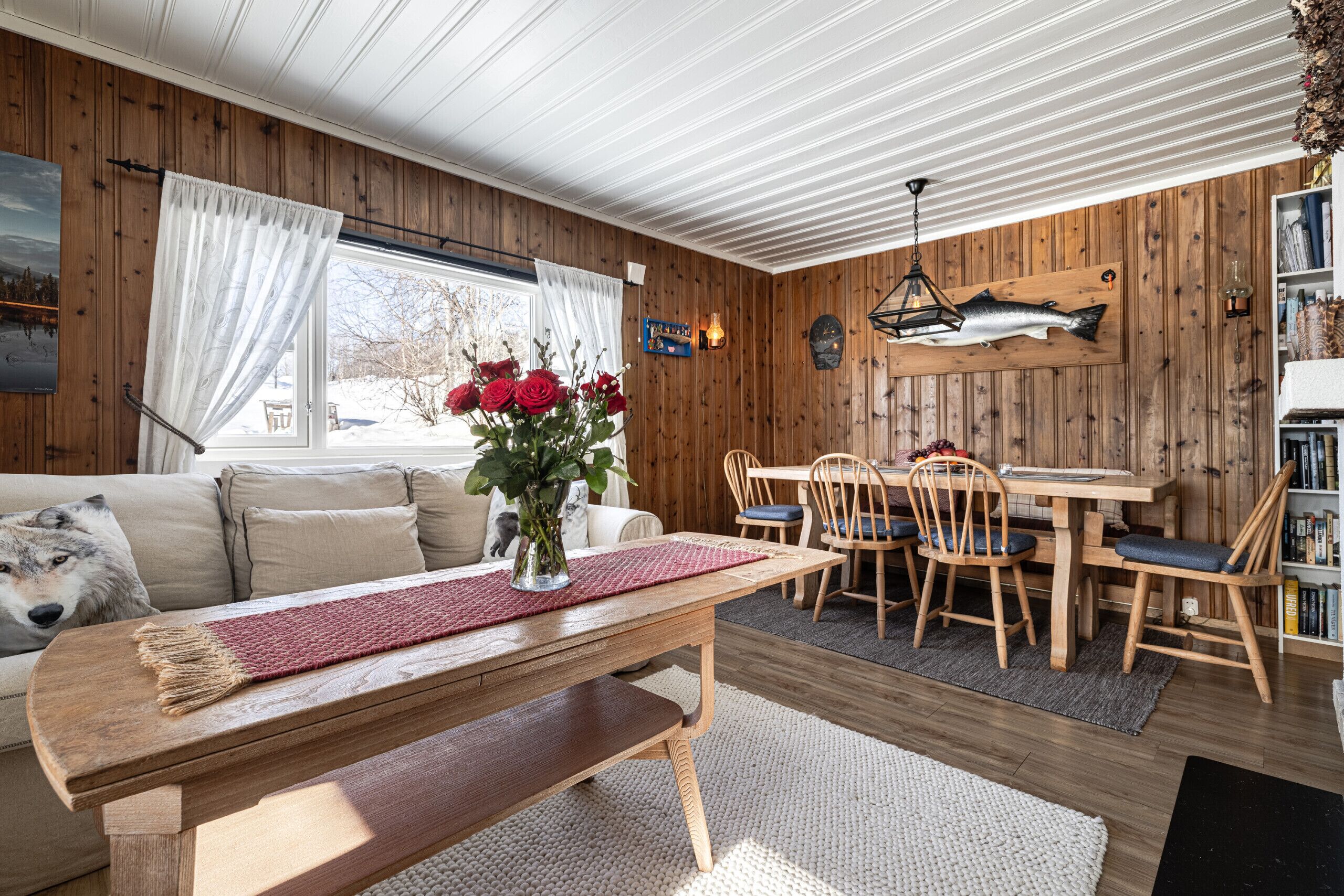 En mysig stuga med en inredning som inkluderar en matplats med träbord, en soffa med ett dekorativt prydnadskudd och en vas med röda blommor.