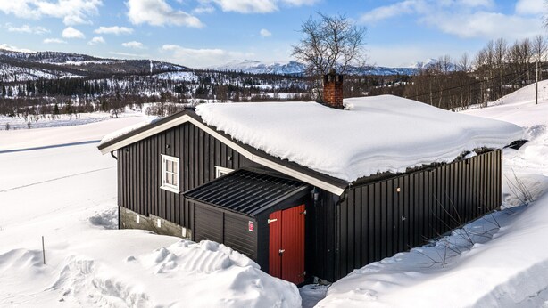 Une petite cabane en bois à un seul étage, avec une porte rouge, entourée de terre enneigée et dominée par un paysage montagneux.