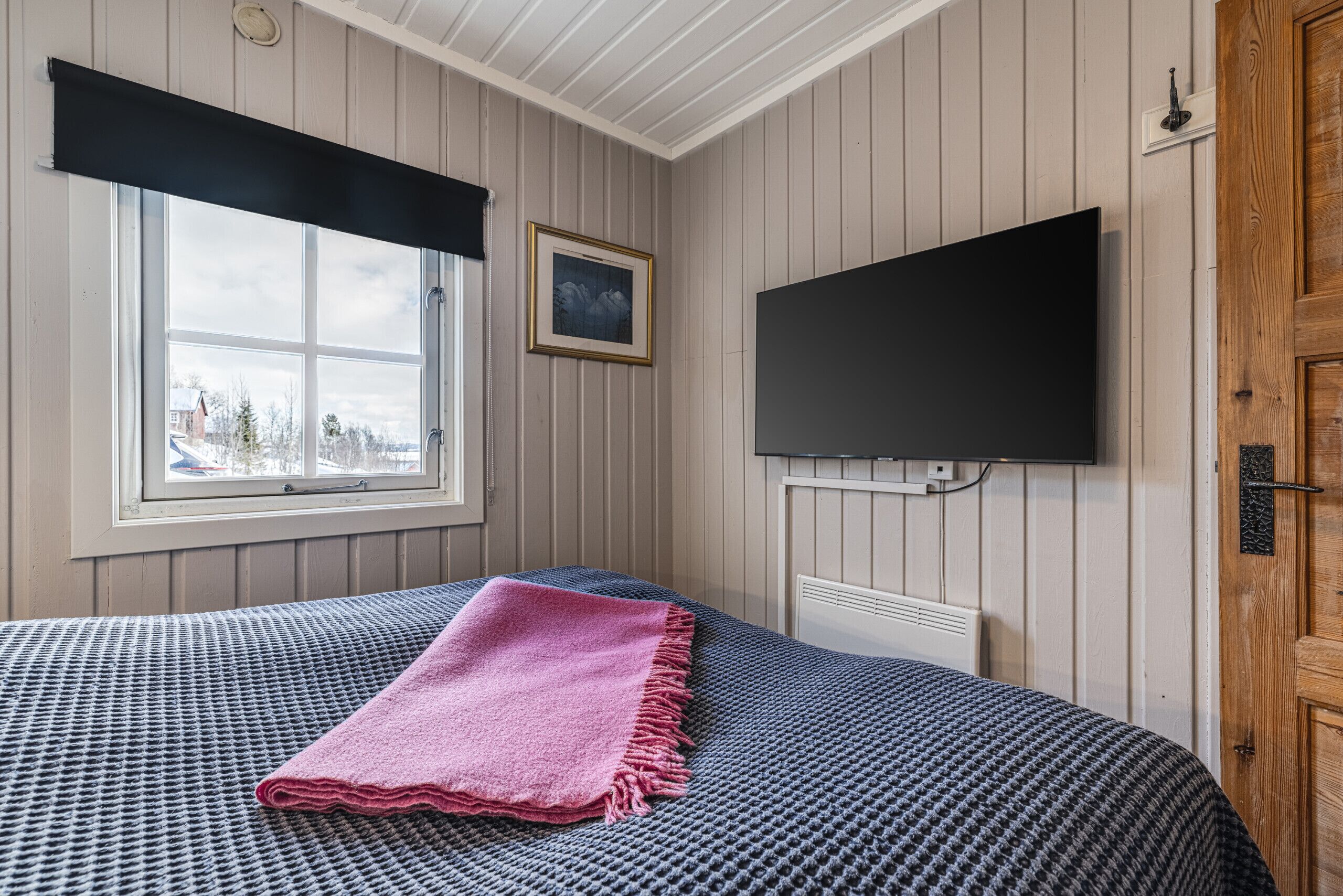 A bedroom with a bed, a window with a view, a mounted TV, and a wooden door.