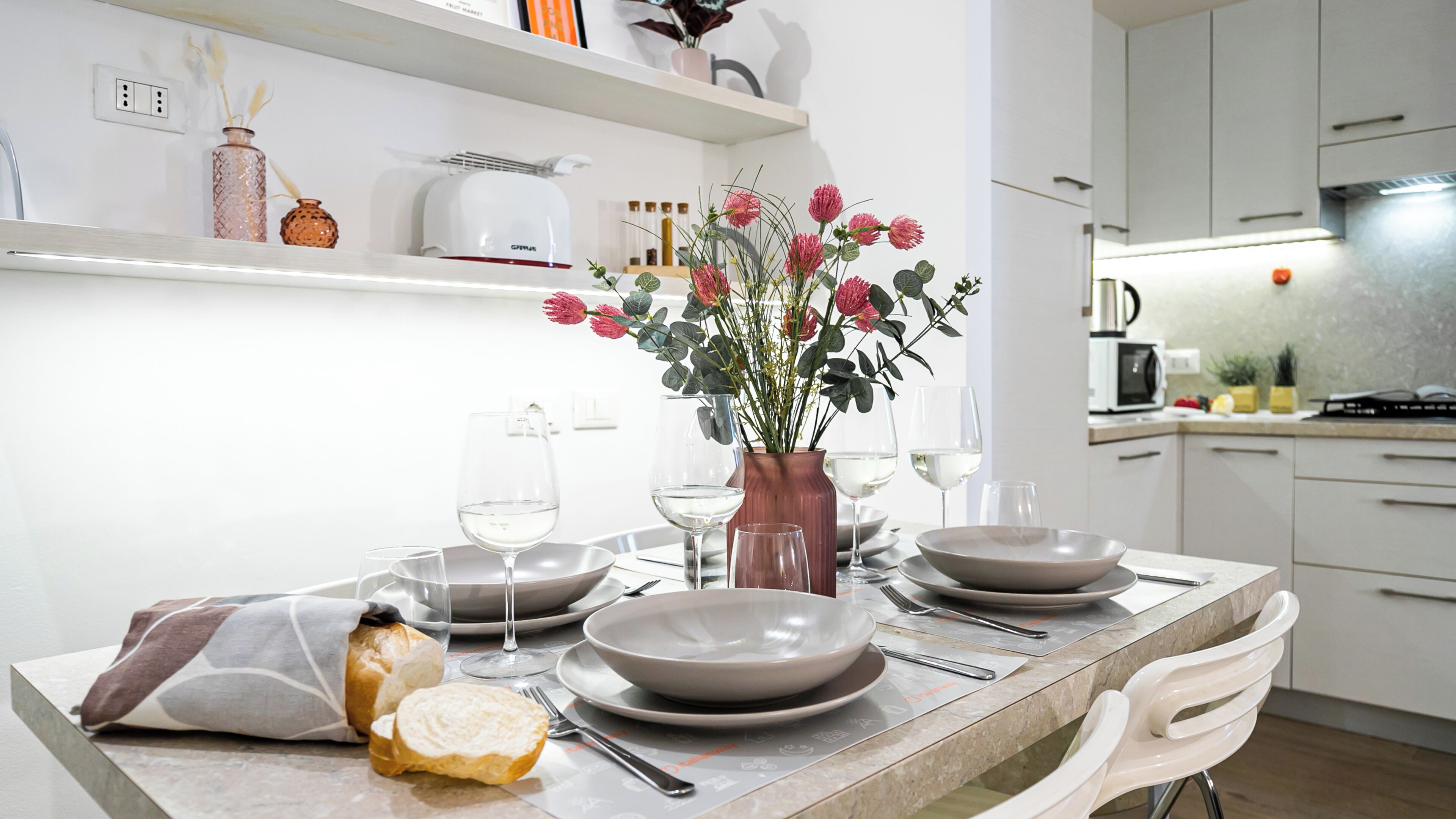 A modern kitchen with white cabinets, a marble countertop, and a dining table set with plates, glasses, and a vase of flowers.