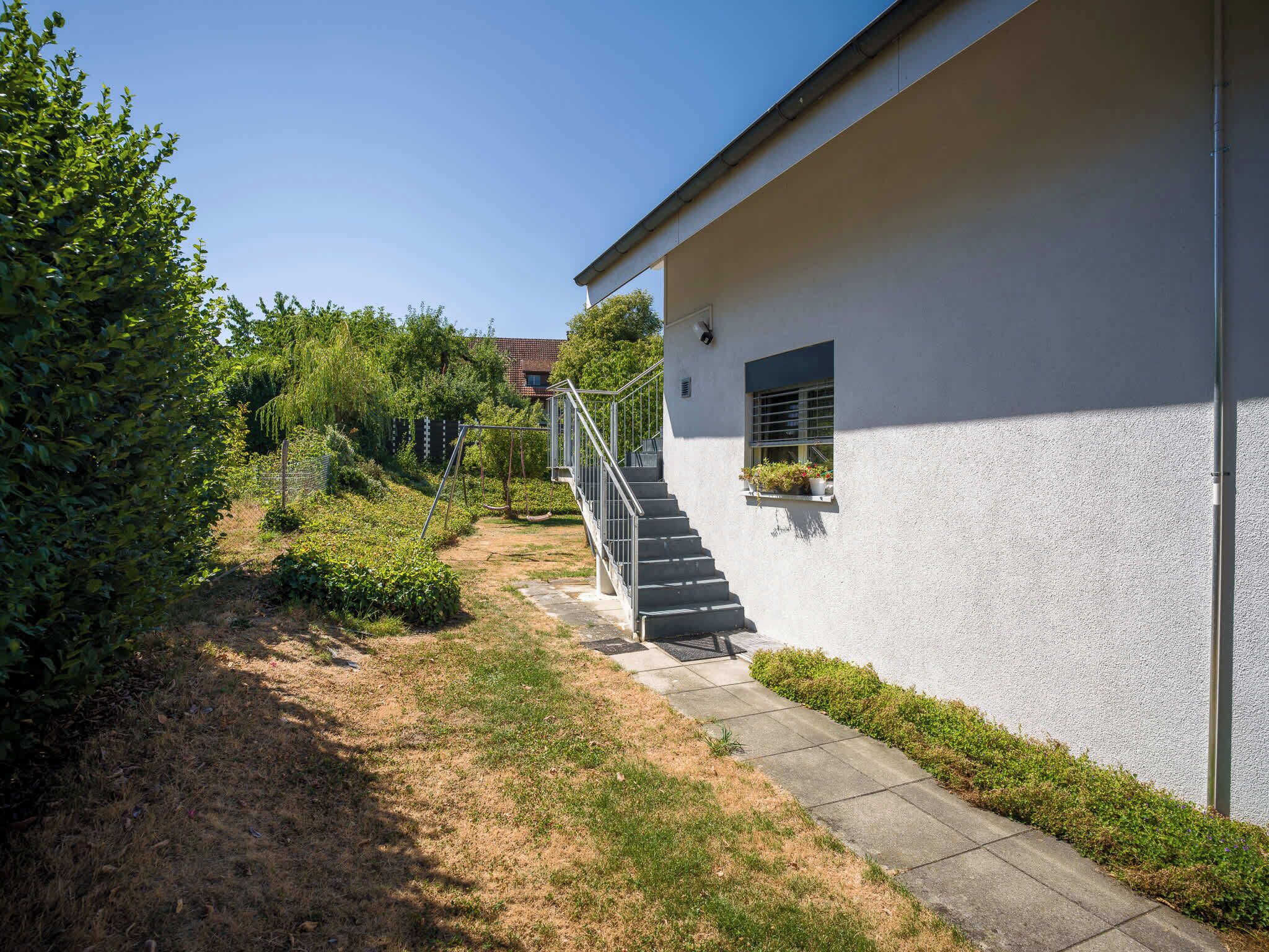 A modern building with a white exterior, a concrete pathway, and a metal staircase leading to a window with potted plants.