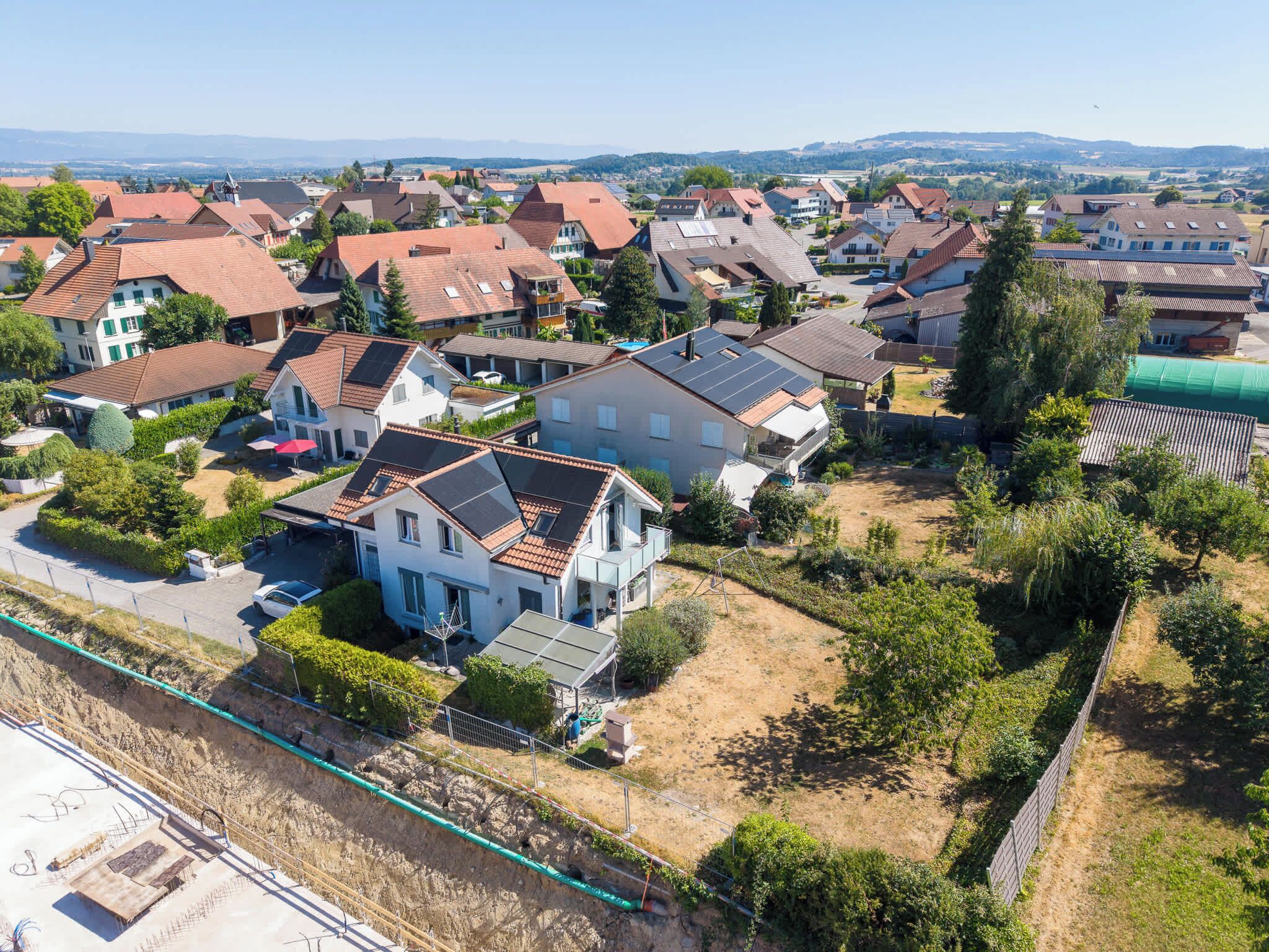 A residential area with houses, some with solar panels, surrounded by greenery and a clear sky.