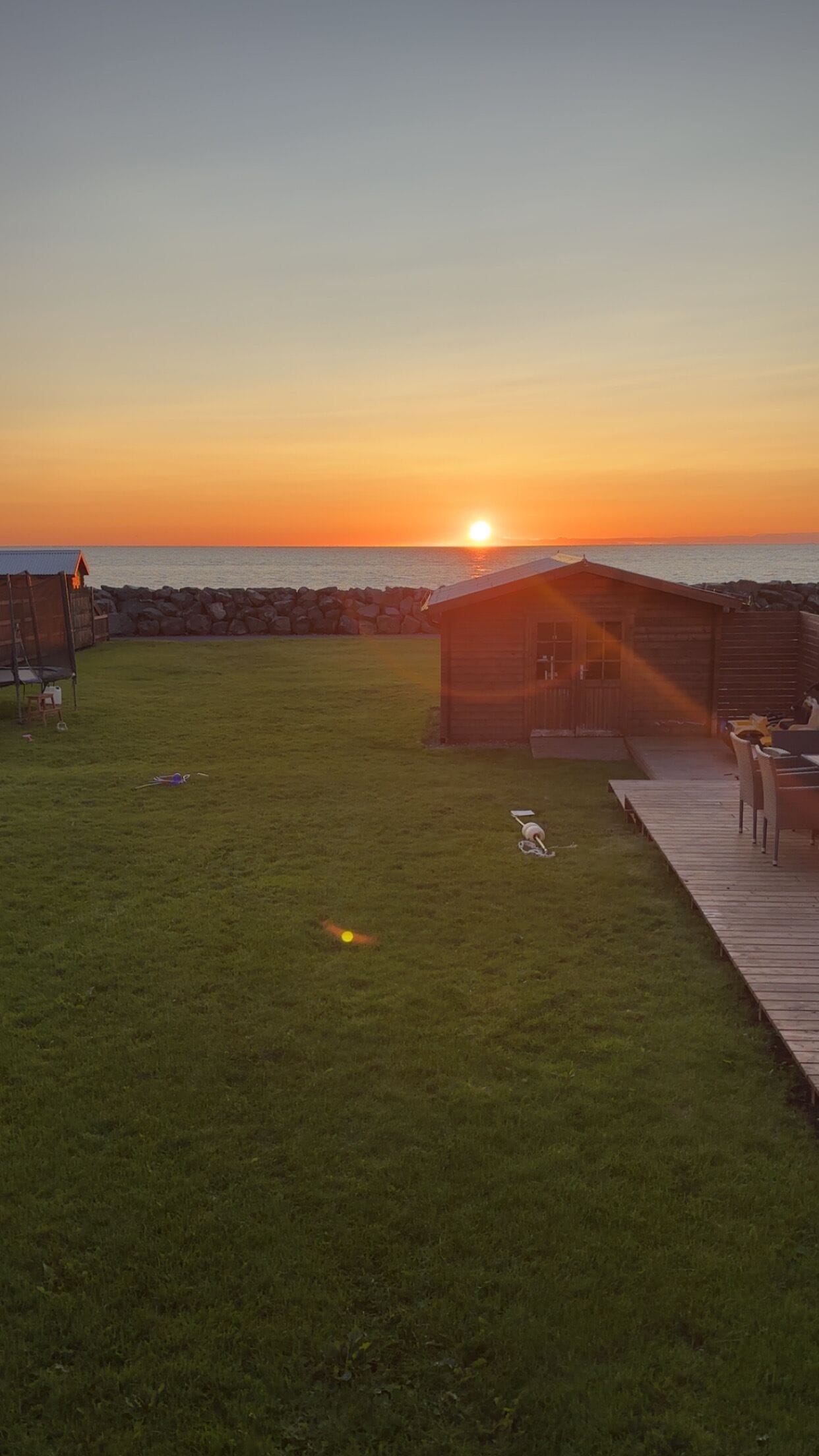 A grassy area with a wooden deck leading to a cabin, a stone wall, and a sunset in the background.