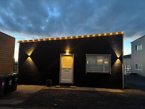 Un petit bâtiment de couleur sombre, doté d’une porte blanche et d’une fenêtre, illuminé par des guirlandes lumineuses.