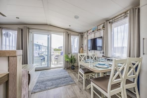 A dining area with a table set for four, a television mounted on the wall, and a view of the outside through a glass door. - Lovely Home in Church Farm Haven Park - Pass the Keys (Pagham)