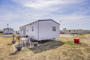 A mobile home with a porch and a small deck, surrounded by dry grass and potted plants. - Lovely Home in Church Farm Haven Park - Pass the Keys (Pagham)