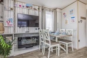 A dining area with a wooden table, white chairs, and a television mounted above the fireplace. - Lovely Home in Church Farm Haven Park - Pass the Keys (Pagham)
