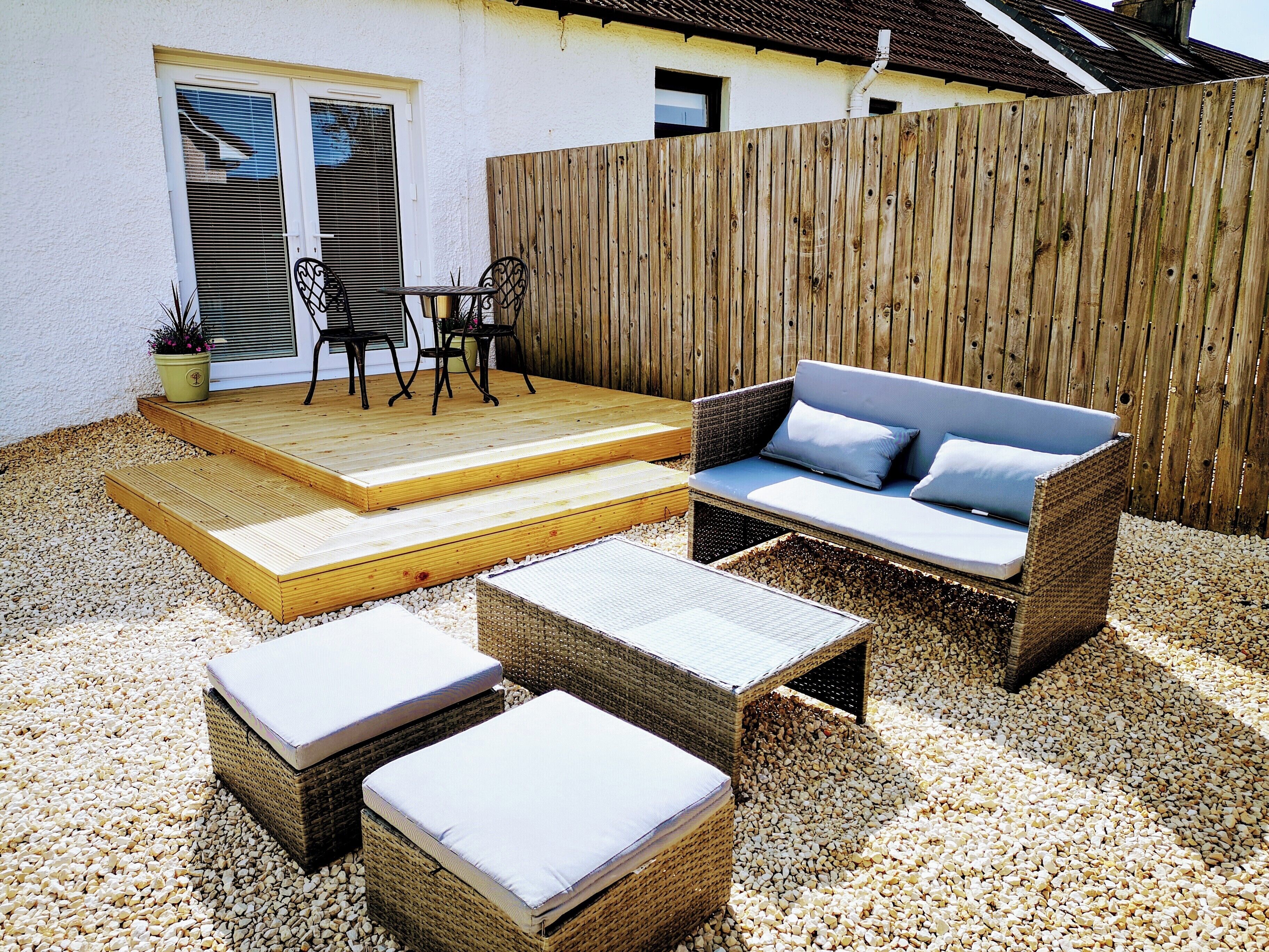 A patio with wicker furniture, a wooden deck, and a white wall with windows.